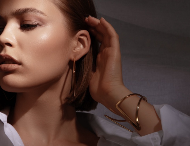 a woman with her eyes closed and her hand against her ear wearing gold jewelry